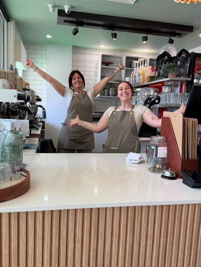 Two smiling women wearing matching brown aprons stand behind a white counter in a cafe, with one raising both arms in a celebratory gesture.