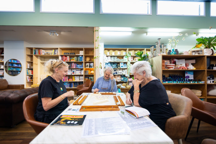 Three women seated around a table in a bright indoor shop or community space, working on a wooden board game. Bookshelves, display shelves, chairs, and windows are visible in the background.