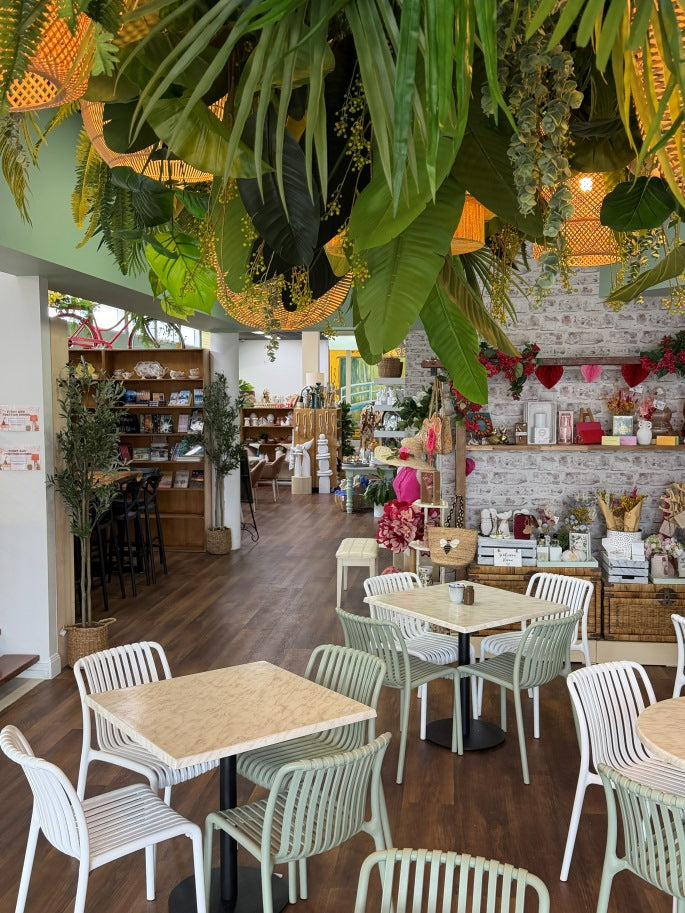 A cozy cafe interior with small square tables and white chairs, surrounded by lush hanging green plants and warm woven pendant lights. The space has dark wood floors, a white brick wall, and shelves with decorative items and flowers.